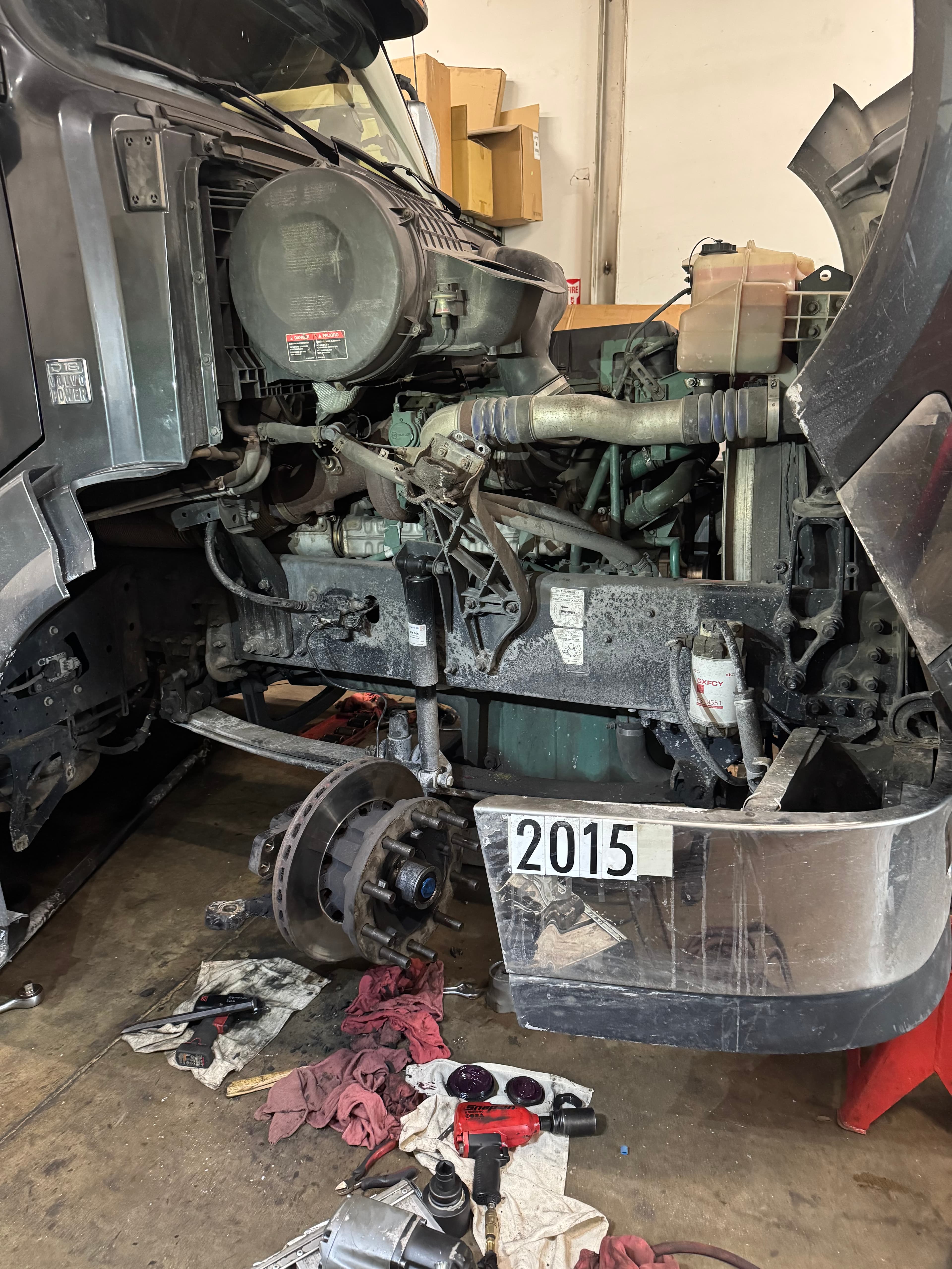 Semi-truck engine bay with open hood and exposed front wheel hub undergoing mechanical repairs.